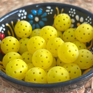 May include: A black ceramic bowl filled with bright yellow pickleballs. The bowl has a decorative floral pattern in blue, white, and red. The pickleballs are round with numerous small holes.