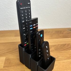 May include: A black remote control organizer with four slots holds four different black remote controls. The organizer is made of plastic and has a modern design.