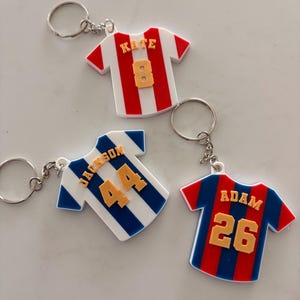 May include: Three keychains shaped like soccer jerseys. One is red and white with "KATE" and the number "8". Another is white and blue with "JACKSON" and "44". The last is red and blue with "ADAM" and "26".