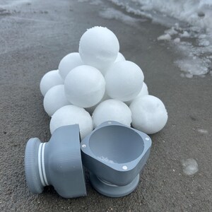 May include: A set of two gray, magnetic, snowball molds. The molds are in the shape of a sphere and a cylinder. The molds are sitting on a gray surface with a pile of white snowballs in the background.