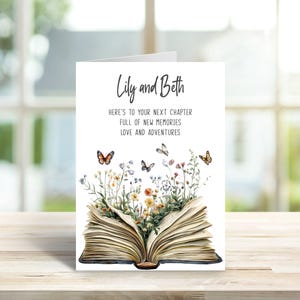 Op de afbeelding: Een witte wenskaart met een open boek waaruit kleurrijke wilde bloemen en vlinders tevoorschijn komen. De tekst in zwarte inkt luidt: Lily and Beth, HERE'S TO YOUR NEXT CHAPTER, FULL OF NEW MEMORIES, LOVE AND ADVENTURES.