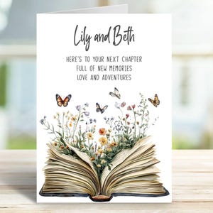 May include: A white greeting card with the names "Lily and Beth" at the top. The card features an open book with watercolor flowers and butterflies. The text reads "HERE'S TO YOUR NEXT CHAPTER FULL OF NEW MEMORIES LOVE AND ADVENTURES."