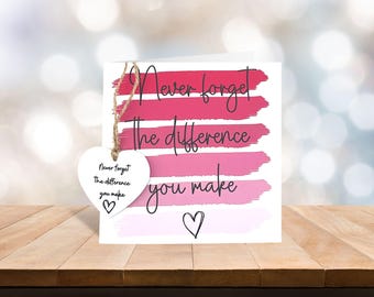 May include: A white greeting card with the message "Never forget the difference you make" in black script. The card features pink brushstrokes and a heart-shaped tag with the same message, tied with twine.