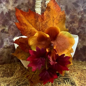 May include: A white basket with a cream-colored handle, adorned with fall-colored leaves and an orange flower. The leaves are in shades of orange, red, and yellow, creating a seasonal theme.