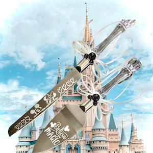 May include: A set of two silver cake cutting knives with black handles and white ribbon bows. The knives are engraved with the words "Believe in Magic" and "Mickey Mouse" and hearts. The knives are in front of a blurred image of Cinderella's Castle.