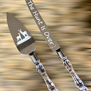 May include: A set of two silver cake knives with crystal handles. The larger knife has the text "The Hunt Is Over" engraved on the blade. The smaller knife has a silhouette of a deer and fawn engraved on the blade.