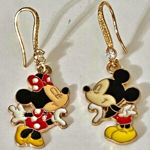 K&ouml;nnte beinhalten: Goldfarbene Ohrh&auml;nger mit Mickey und Minnie Mouse. Die Ohrringe haben einen goldenen Haken mit einem kleinen, klaren Edelstein. Minnie Mouse tr&auml;gt ein rotes Polka-Dot-Kleid und eine Schleife. Mickey Mouse tr&auml;gt rote Shorts und gelbe Schuhe.