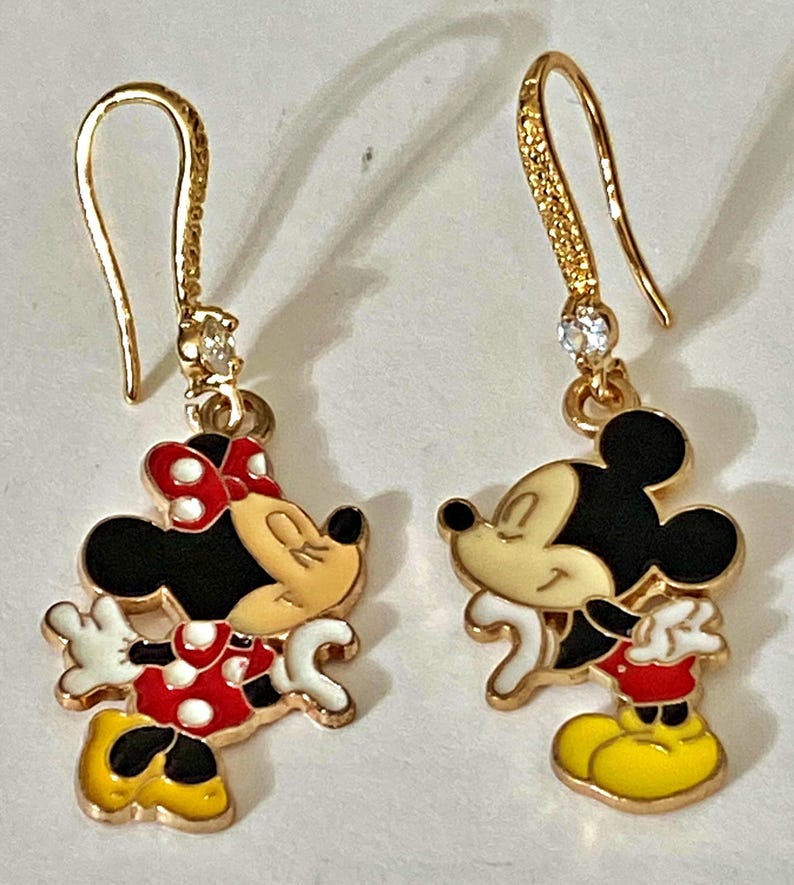K&ouml;nnte beinhalten: Goldfarbene Ohrh&auml;nger mit Mickey und Minnie Mouse. Die Ohrringe haben einen goldenen Haken mit einem kleinen, klaren Edelstein. Minnie Mouse tr&auml;gt ein rotes Polka-Dot-Kleid und eine Schleife. Mickey Mouse tr&auml;gt rote Shorts und gelbe Schuhe.