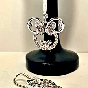 May include: Silver-coloured dangle earrings with a Minnie Mouse design. The earrings feature a bow and are encrusted with clear crystals. One earring is displayed on a black stand, the other lies on a white surface.