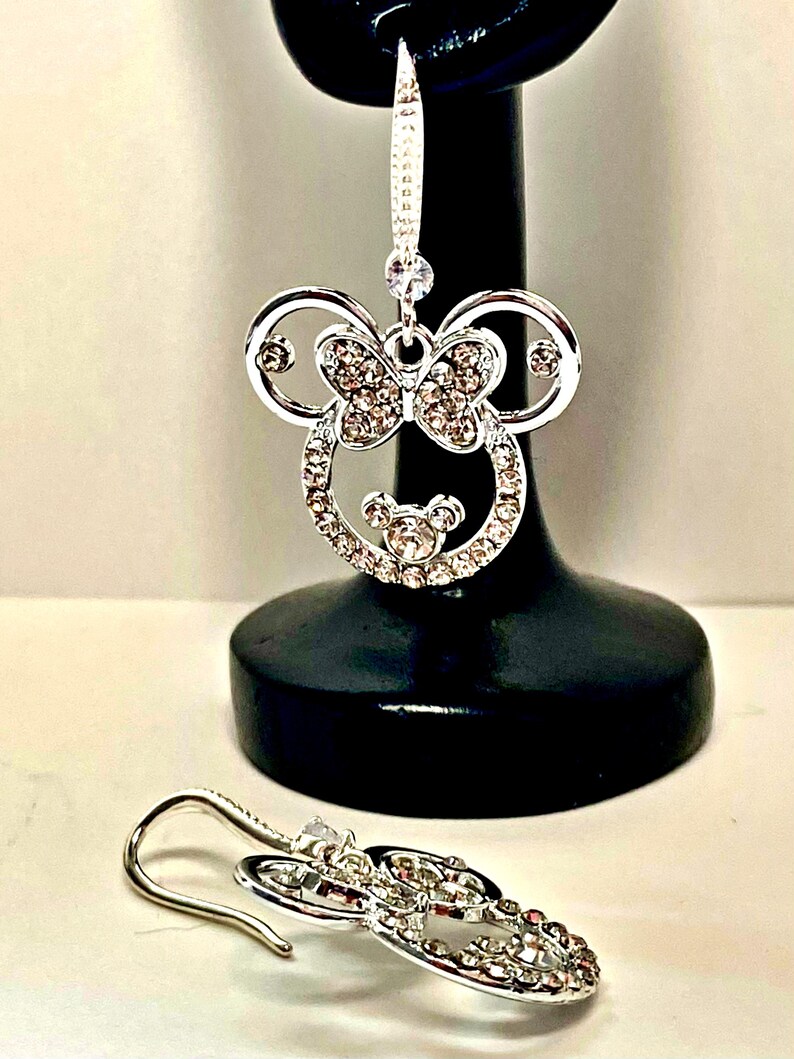 May include: Silver-coloured dangle earrings with a Minnie Mouse design. The earrings feature a bow and are encrusted with clear crystals. One earring is displayed on a black stand, the other lies on a white surface.