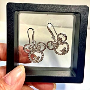 May include: A pair of silver-coloured earrings shaped like a mouse head with a bow, studded with small crystals. The earrings are presented in a black display box.