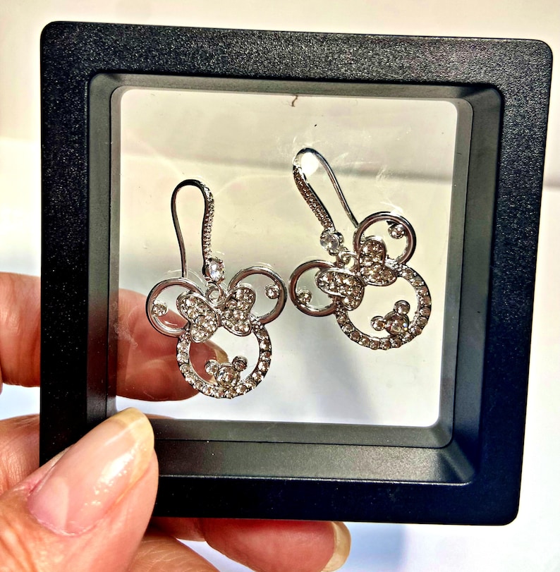 May include: A pair of silver-coloured earrings shaped like a mouse head with a bow, studded with small crystals. The earrings are presented in a black display box.