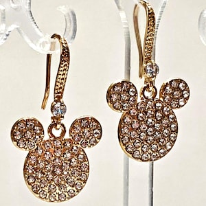May include: Gold-toned dangle earrings featuring a Mickey Mouse design. Each earring has a large, round head and two smaller circles for ears, all encrusted with small, sparkling crystals. The earrings hang from a textured hook.