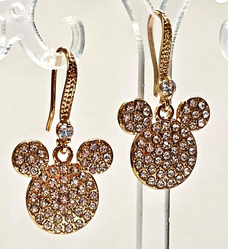 May include: Gold-toned dangle earrings featuring a Mickey Mouse design. Each earring has a large, round head and two smaller circles for ears, all encrusted with small, sparkling crystals. The earrings hang from a textured hook.