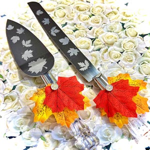 May include: A set of two silver cake knives with fall leaf designs and red and yellow fall leaf accents. The knives are on a white background.