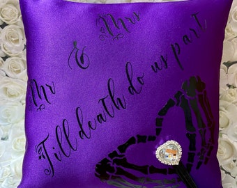 May include: A square purple satin ring pillow featuring black script text that reads Mr & Mrs and Till death do us part. A black graphic of two skeletal hands forming a heart shape is printed on the fabric, accented by a heart-shaped rhinestone charm.