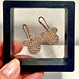 May include: A pair of gold-colored earrings in the shape of Mickey Mouse's head. The earrings are covered in small, sparkling crystals. The earrings are displayed in a black box with a white background.