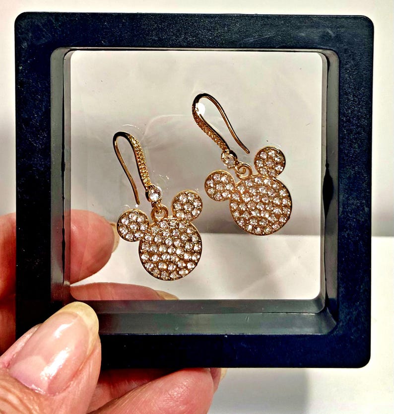 May include: A pair of gold-colored earrings in the shape of Mickey Mouse's head. The earrings are covered in small, sparkling crystals. The earrings are displayed in a black box with a white background.