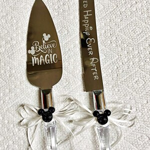 May include: A set of two silver cake knives with black Mickey Mouse ears on the handles. The knife on the left has the words "Believe in Magic" engraved on the blade. The knife on the right has the words "They Lived Happily Ever After" engraved on the blade.