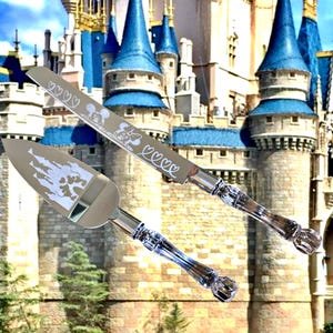 May include: Silver cake knife and server set with clear crystal handles. The knife blade features etched Mickey and Minnie Mouse designs with hearts. The server has a castle and character design. The background is a castle.