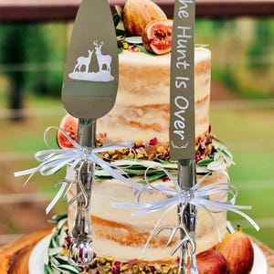 May include: A wedding cake cutting set with two silver knives. One knife has a deer silhouette and the other knife has the text "The Hunt Is Over". The knives are tied with white ribbon and are on a two-tiered cake decorated with figs and greenery.