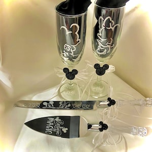 May include: A wedding cake cutting set with a black and silver knife and server. The server has the text "Believe in Magic" engraved on it. The handles of the knife and server are decorated with black Mickey Mouse ears. Two champagne flutes with black Mickey Mouse ears are also included.