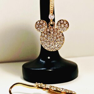 May include: Gold-toned Mickey Mouse-shaped earrings with sparkling crystal accents. The earrings feature a classic three-circle design, with the main circle and two smaller circles covered in crystals. One earring is displayed on a black stand, and the other lies on a white surface.