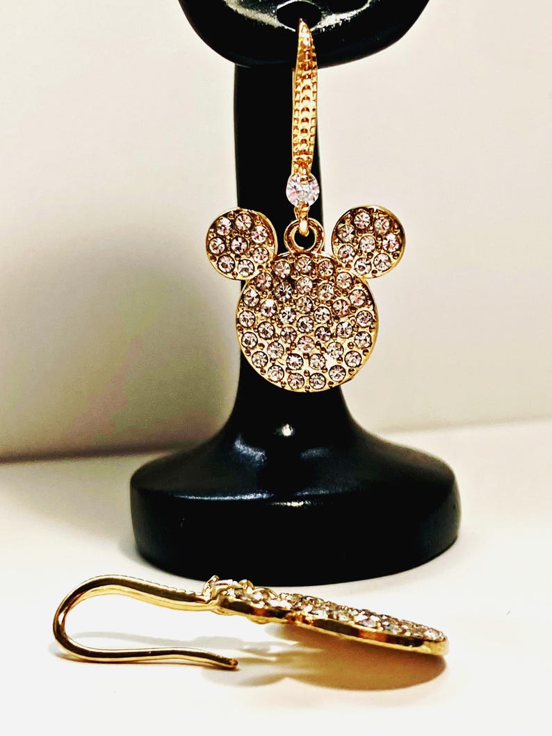 May include: Gold-toned Mickey Mouse-shaped earrings with sparkling crystal accents. The earrings feature a classic three-circle design, with the main circle and two smaller circles covered in crystals. One earring is displayed on a black stand, and the other lies on a white surface.