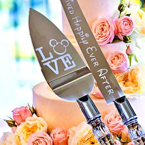 May include: Silver cake knife and server set with the words "They Lived Happily Ever After" and "Love" with a Mickey Mouse head. The set is displayed in front of a multi-tiered cake decorated with pink and yellow roses.