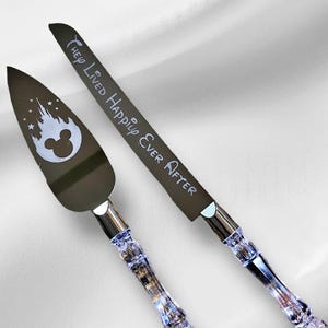 May include: A silver cake knife and server set with clear crystal handles. The knife has the inscription "They Lived Happily Ever After" and the server has a silhouette of the Disney castle and Mickey Mouse head.