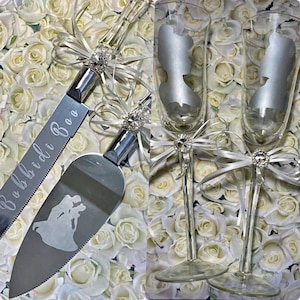May include: A wedding cake cutting set with a silver knife and server. The knife has the words "Bibbidi Boo" engraved on it. The server has a silhouette of a couple dancing. Two champagne flutes with a silhouette of a woman on each glass are included. The set is decorated with white satin ribbons and rhinestones.