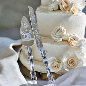 May include: Silver cake knife and server set with clear, faceted handles. The server features a castle design and decorative swirls. The knife and server are displayed in front of a white tiered cake decorated with white roses.