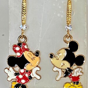 K&ouml;nnte beinhalten: Ein Paar goldfarbene Ohrh&auml;nger mit Mickey und Minnie Mouse. Jeder Ohrring hat einen goldfarbenen Reifen mit einem kleinen, klaren Edelstein. Die Mickey- und Minnie Mouse-Figuren sind in ihren klassischen Outfits mit schwarzen, roten, gelben und wei&szlig;en Details.