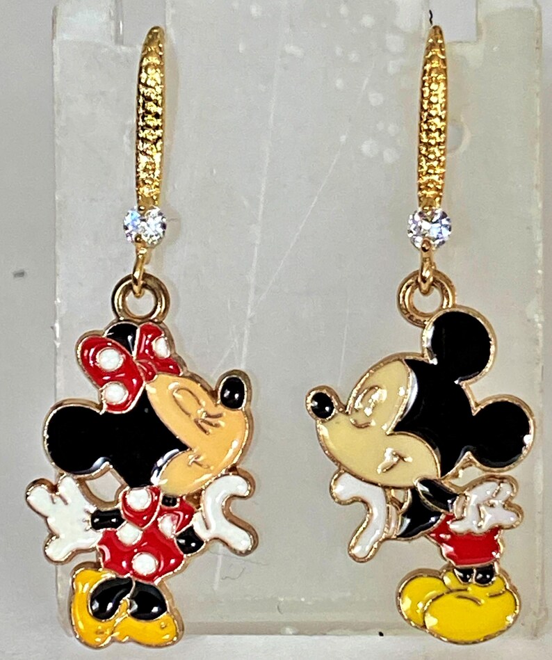 K&ouml;nnte beinhalten: Ein Paar goldfarbene Ohrh&auml;nger mit Mickey und Minnie Mouse. Jeder Ohrring hat einen goldfarbenen Reifen mit einem kleinen, klaren Edelstein. Die Mickey- und Minnie Mouse-Figuren sind in ihren klassischen Outfits mit schwarzen, roten, gelben und wei&szlig;en Details.