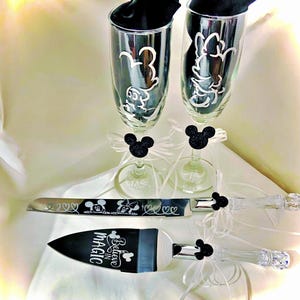 May include: A wedding set featuring two champagne flutes with Mickey and Minnie Mouse designs, a cake knife, and a server. The flutes have black Mickey Mouse embellishments and white ribbons. The cake knife and server have clear handles.