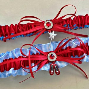 May include: Two ruffled garters, one red and one blue, with red bows and rhinestone accents. One garter has a silver star wand charm, the other has red shoe charms. The garters are approximately 1.5 inches wide.