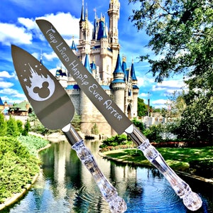 May include: A set of two silver cake servers with clear plastic handles. The server on the left has a Mickey Mouse head and a silhouette of Cinderella's castle. The server on the right has the text "They Lived Happily Ever After". The servers are in front of Cinderella's castle at Walt Disney World.