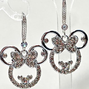 May include: Silver-coloured drop earrings with a Minnie Mouse design. Each earring features a circular head shape with a bow and small round face, all encrusted with sparkling crystals. The earrings hang from a decorative lever back.
