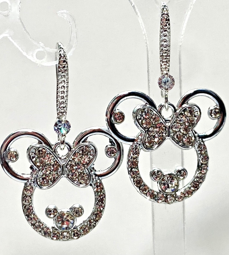 May include: Silver-coloured drop earrings with a Minnie Mouse design. Each earring features a circular head shape with a bow and small round face, all encrusted with sparkling crystals. The earrings hang from a decorative lever back.