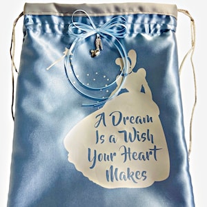 May include: A light blue satin drawstring bag with a white silhouette of Cinderella and the text "A Dream Is a Wish Your Heart Makes". A blue ribbon with a silver glass slipper charm is tied to the top of the bag.