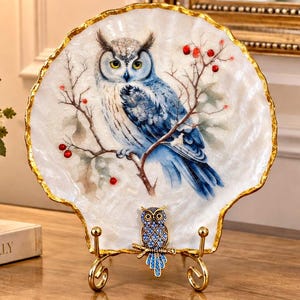 May include: A decorative shell-shaped plate with a white pearlescent finish and a gold-painted scalloped edge. The center features a detailed illustration of a blue and white owl perched on a branch with red berries. A small blue owl ornament sits on the gold metal display stand.