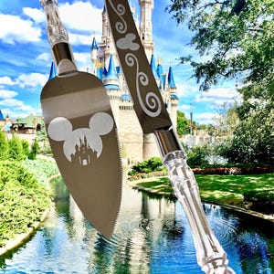 May include: A silver cake knife and server with clear plastic handles. The knife has a swirl design and a Mickey Mouse head. The server has a Mickey Mouse head with the Cinderella Castle silhouette. The background is a blurred image of the Cinderella Castle at Walt Disney World.