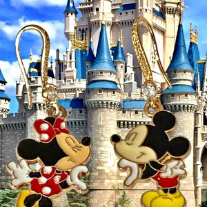 K&ouml;nnte beinhalten: Goldfarbene Mickey Mouse und Minnie Mouse Ohrh&auml;nger. Minnie tr&auml;gt ein rot-wei&szlig; gepunktetes Kleid und eine Schleife. Mickey tr&auml;gt rote Shorts und gelbe Schuhe. Im Hintergrund ist ein Schloss zu sehen.