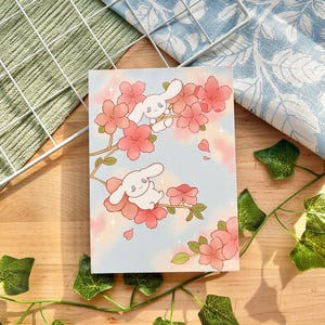 May include: A postcard with a cartoon illustration of two white bunny-like characters amidst pink cherry blossoms on a light blue backdrop. The postcard rests on a wooden surface, with green ivy and a white wire grid.