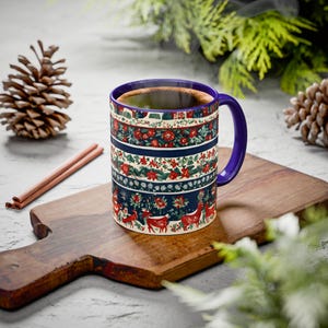 May include: A ceramic mug with a blue handle and rim, filled with a dark liquid. The mug features a festive design with red flowers, green leaves, and reindeer on a navy blue background. The mug is placed on a wooden cutting board.