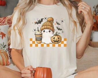 Puede incluir: Camiseta color crema con un fantasma de dibujos animados que lleva un gorro de punto, sosteniendo un café con leche de calabaza y junto a una calabaza tallada. El diseño incluye murciélagos y estrellas, con el texto "Pumpkin Spice".