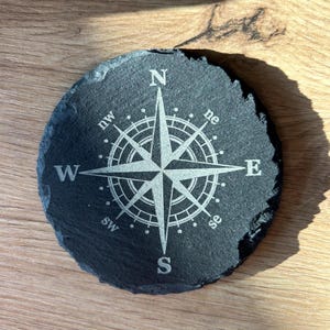May include: A black slate coaster featuring a white compass design. The compass points north, south, east, and west, with the letters N, S, E, and W. The compass also includes the points NW, NE, SW, and SE.