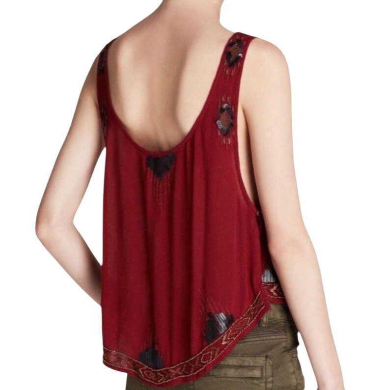 May include: Burgundy tank top with a low scoop back and embellished straps. The top has black and gold beaded accents on the straps and hemline. The fabric looks lightweight and flowing, suitable for warm weather.