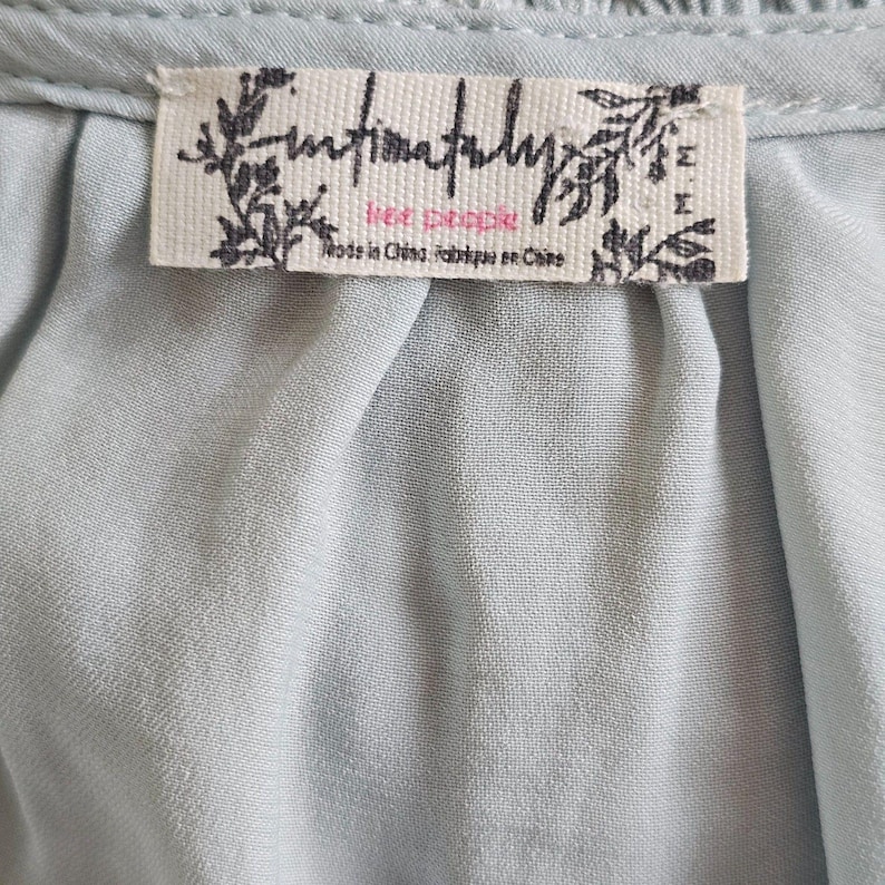 Może przedstawiać: Zbliżenie jasnoniebieskiej tkaniny z metką. Na metce widnieje napis "Intimately Free People" z tekstem poniżej "Made in China/Fabriqu&eacute; en Chine". Tkanina jest lekko pomarszczona.
