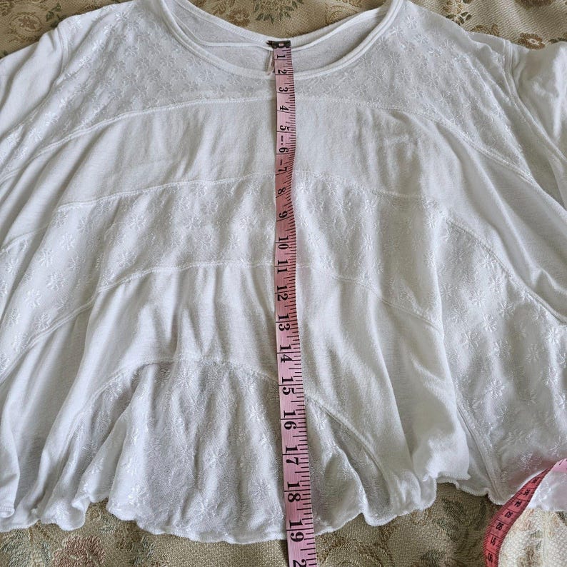 以下が含まれることがあります： 白いフローイングトップ、花の刺繍とラウンドネックが特徴です。 ピンクのメジャーで測定すると、長さは約48cmです。 生地は柔らかく、軽量に見えます。